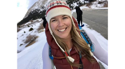 A woman in a winter hat smiles at the camera as she stands on a snowy winter road