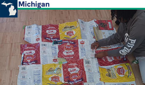 Student working on creating insulated sleeping mat out of chip bags and wrappers; small map of Michigan
