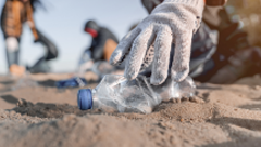 Gloved hand picking up plastic bottle from sandy ground