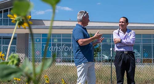 Two people standing in front of fence and talking with yellow flowers in foreground