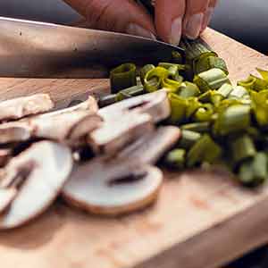 Close up of person chopping mushrooms and green onions with knife on cutting board